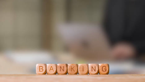 Business-Concept-Wooden-Letter-Cubes-Or-Dice-Spelling-Banking-With-Office-Person-Working-At-Laptop-In-Background