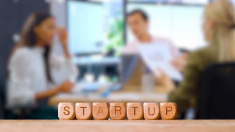 Business-Concept-Wooden-Letter-Cubes-Or-Dice-Spelling-Startup-With-Office-Meeting-In-Background