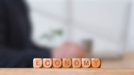 Business-Concept-Wooden-Letter-Cubes-Or-Dice-Spelling-Economy-With-Office-Person-Using-Mobile-Phone-In-Background