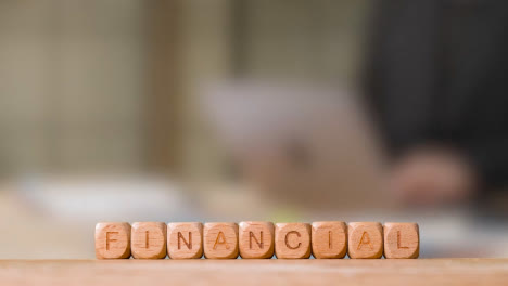 Business-Concept-Wooden-Letter-Cubes-Or-Dice-Spelling-Financial-With-Person-Working-On-Laptop-In-Office-In-Background