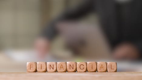 Business-Concept-Wooden-Letter-Cubes-Or-Dice-Spelling-Financial-With-Person-Working-On-Laptop-In-Office-In-Background