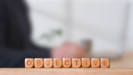 Business-Concept-Wooden-Letter-Cubes-Or-Dice-Spelling-Objective-With-Office-Person-Using-Mobile-Phone-In-Background