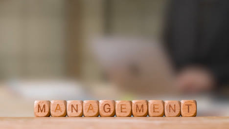 Business-Concept-Wooden-Letter-Cubes-Or-Dice-Spelling-Management-With-Person-Working-On-Laptop-In-Office-In-Background