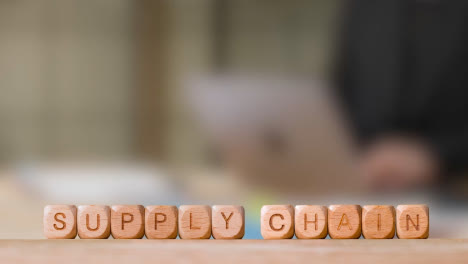 Business-Concept-Wooden-Letter-Cubes-Or-Dice-Spelling-Supply-Chain-With-Person-Working-On-Laptop-In-Office-In-Background