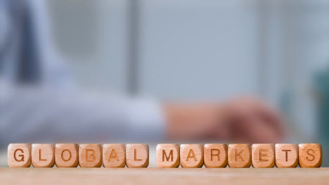 Business-Concept-Wooden-Letter-Cubes-Or-Dice-Spelling-Global-Markets-With-Office-Person-Working-At-Laptop-In-Background