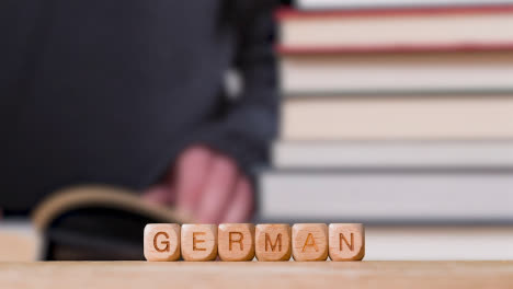 Education-Concept-Shot-With-Wooden-Letter-Cubes-Or-Dice-Spelling-German-With-Person-Reading-Book-In-Library