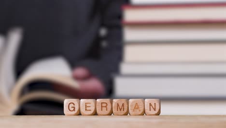 Education-Concept-Shot-With-Wooden-Letter-Cubes-Or-Dice-Spelling-German-With-Person-Reading-Book-In-Library