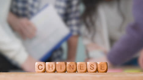 Education-Concept-With-Wooden-Letter-Cubes-Or-Dice-Spelling-Chinese-With-Students-Meeting-In-Background