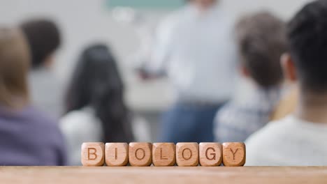 Education-Concept-With-Wooden-Letter-Cubes-Or-Dice-Spelling-Biology-With-Student-Lecture-In-Background
