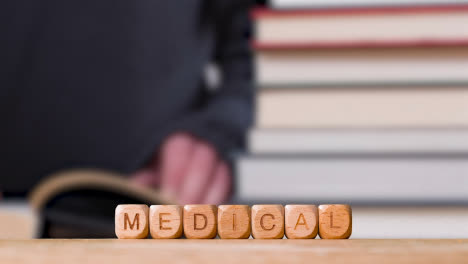 Education-Concept-Shot-With-Wooden-Letter-Cubes-Or-Dice-Spelling-Medical-With-Person-Reading-Book-In-Library