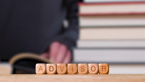 Education-Concept-Shot-With-Wooden-Letter-Cubes-Or-Dice-Spelling-Advisor-With-Person-Reading-Book-In-Library