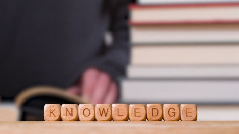 Education-Concept-Shot-With-Wooden-Letter-Cubes-Or-Dice-Spelling-Knowledge-With-Person-Reading-Book-In-Library