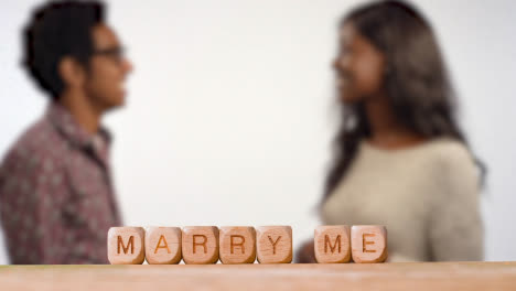 Concept-With-Wooden-Letter-Cubes-Or-Dice-Spelling-Marry-Me-Against-Background-Of-Couple-Hugging