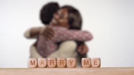 Concept-With-Wooden-Letter-Cubes-Or-Dice-Spelling-Marry-Me-Against-Background-Of-Couple-Hugging