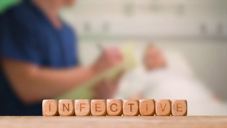 Medical-Concept-With-Wooden-Letter-Cubes-Or-Dice-Spelling-Infective-Against-Background-Of-Nurse-Talking-To-Patient-In-Hospital-Bed