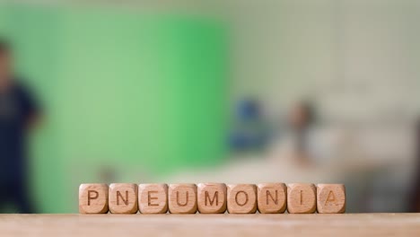 Medical-Concept-With-Wooden-Letter-Cubes-Or-Dice-Spelling-Pneumonia-Against-Background-Of-Nurse-Talking-To-Patient-In-Hospital-Bed