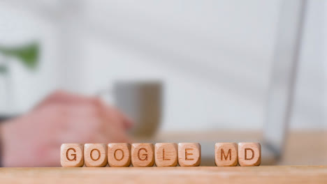 Medical-Concept-With-Wooden-Letter-Cubes-Or-Dice-Spelling-Google-MD-Against-Background-Of-Man-Looking-At-Laptop