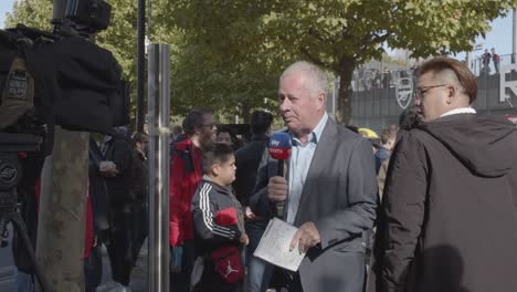 TV-Sports-Reporter-Outside-The-Emirates-Stadium-Home-Ground-Arsenal-Football-Club-London-With-Supporters-On-Match-Day