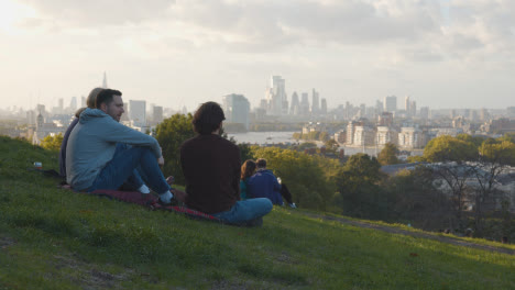 People-Sitting-On-Hill-In-Greenwich-Park-Looking-Out-Over-View-Of-City-Skyline-And-River-Thames-In-London-UK