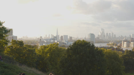 People-Sitting-On-Hill-In-Greenwich-Park-Looking-Out-Over-View-Of-City-Skyline-And-River-Thames-In-London-UK-1
