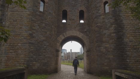 Person-Walking-Through-Gate-In-Castle-Walls-In-Bute-Park-In-Cardiff-Wales-With-City-Skyline