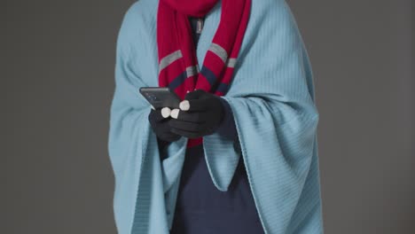 Close-Up-Of-Person-Wearing-Blanket-And-Gloves-Using-Mobile-Phone-Trying-To-Keep-Warm-In-Energy-Crisis