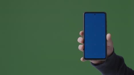 Close-Up-Of-Person-Holding-Blue-Screen-Mobile-Phone-Against-Green-Background