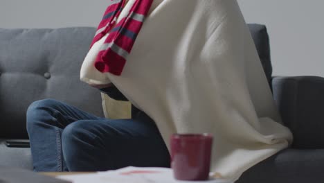 Person-Wearing-Blanket-With-Hot-Drink-Sitting-On-Sofa-At-Home-Trying-To-Keep-Warm-In-Energy-Crisis-2
