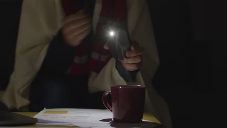Person-Wearing-Blanket-Sitting-On-Sofa-At-Home-In-Dark-When-Power-Is-Cut-Off-Using-Phone-Torch-2