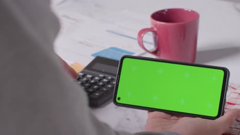 Close-Up-Of-Person-Holding-Green-Screen-Mobile-Phone-Looking-Through-Bills-In-Kitchen-2