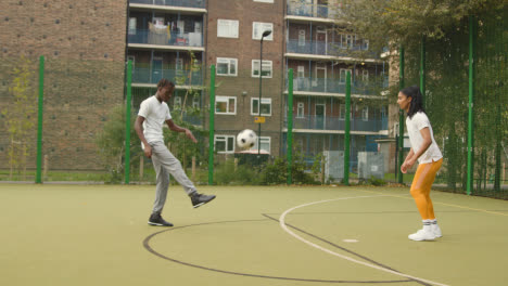Young-Couple-Kicking-Football-On-Artificial-Soccer-Pitch-In-Urban-City-Area-