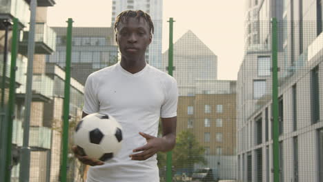 Portrait-Of-Young-Man-With-Football-Under-Arm-On-Artificial-Soccer-Pitch-In-Urban-City-Area-