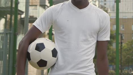 Young-Man-With-Football-Under-Arm-On-Artificial-Soccer-Pitch-In-Urban-City-Area-