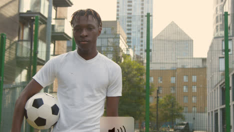 Portrait-Of-Young-Man-With-Football-Under-Arm-On-Artificial-Soccer-Pitch-In-Urban-City-Area-1