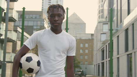 Portrait-Of-Young-Man-With-Football-Under-Arm-On-Artificial-Soccer-Pitch-In-Urban-City-Area-1