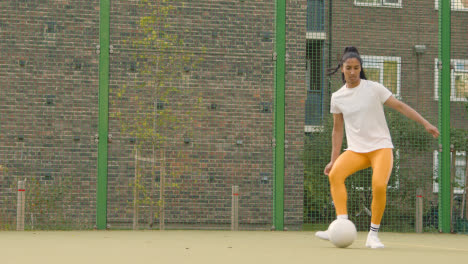 Female-Player-Kicking-And-Passing-Football-On-Artificial-Soccer-Pitch-In-Urban-City-Area-