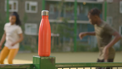 Players-Football-Training-On-Artificial-Soccer-Pitch-In-Urban-City-Area-With-Water-Bottle-In-Foreground