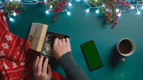 Overhead-Shot-Of-Person-With-Green-Screen-Mobile-Phone-With-Christmas-Decorations-And-Gift-Of-Perfume