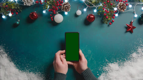 Overhead-Shot-Of-Person-Holding-Green-Screen-Mobile-Phone-Above-Christmas-Decorations