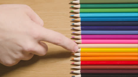 Coloured-Pencils-Arranged-In-A-Line-On-Wooden-Background-With-Finger-Pointing-To-Yellow