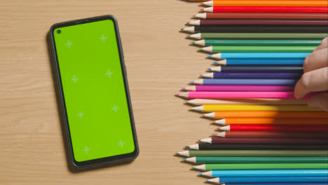 Coloured-Pencils-In-A-Line-On-Wooden-Background-With-Person-Replacing-Pink-One-And-Green-Screen-Mobile-Phone