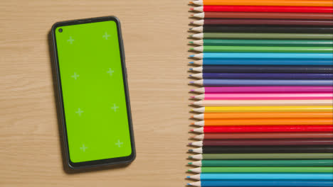 Coloured-Pencils-In-A-Line-On-Wooden-Background-With-Person-Scrolling-On-Green-Screen-Mobile-Phone
