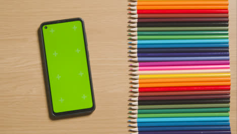 Coloured-Pencils-In-A-Line-On-Wooden-Background-With-Person-Scrolling-On-Green-Screen-Mobile-Phone-1