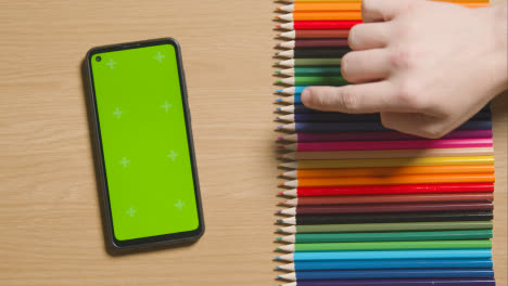 Coloured-Pencils-In-A-Line-On-Wooden-Background-With-Person-Choosing-Purple-One-And-Green-Screen-Mobile-Phone