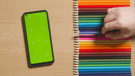 Coloured-Pencils-In-A-Line-On-Wooden-Background-With-Person-Replacing-Purple-One-And-Green-Screen-Mobile-Phone
