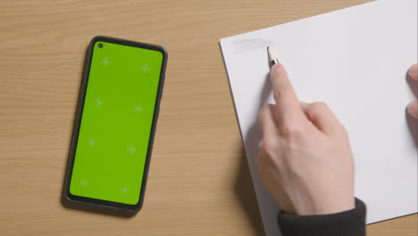 Person-Sketching-With-Pencil-On-Pad-On-Wooden-Table-Background-Next-To-Green-Screen-Mobile-Phone-