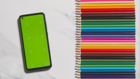 Coloured-Pencils-In-A-Line-On-Marble-Background-With-Person-Scrolling-Through-Green-Screen-Mobile-Phone