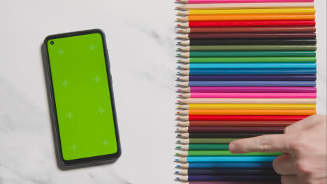 Coloured-Pencils-In-A-Line-On-Marble-Background-With-Person-Choosing-Red-One-And-Green-Screen-Mobile-Phone