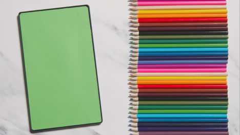Lápices-De-Colores-En-Una-Línea-Sobre-Fondo-De-Mármol-Con-Una-Persona-Desplazándose-Por-Una-Tableta-Digital-De-Pantalla-Verde