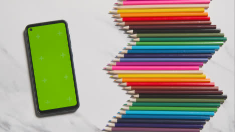 Lápices-De-Colores-Dispuestos-En-Un-Patrón-Simétrico-Con-Teléfono-Móvil-De-Pantalla-Verde-Sobre-Fondo-De-Mármol-Blanco